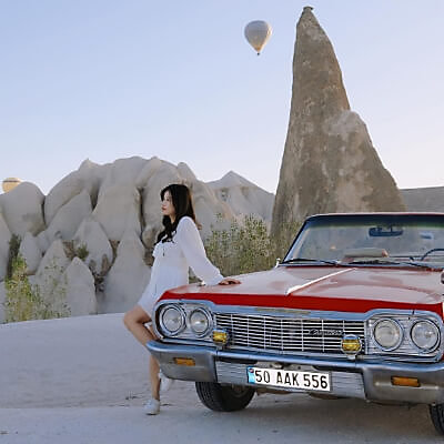 A person wearing a pair of Keds Champion Originals in white, leaning against a classic car in the desert with a hotair balloon high above in the sky.